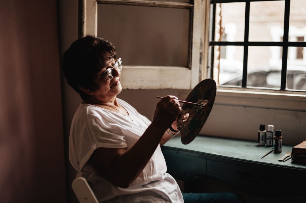A mature woman with glasses paints by a window, creating art in natural light.