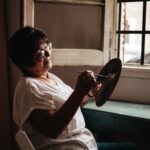 A mature woman with glasses paints by a window, creating art in natural light.