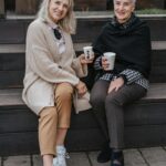 Two senior women sit on a bench outdoors, enjoying coffee and smiling at the camera.