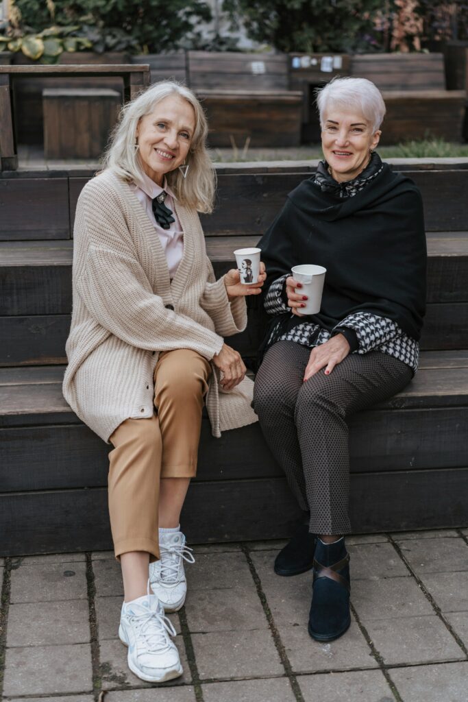 Two senior women sit on a bench outdoors, enjoying coffee and smiling at the camera.