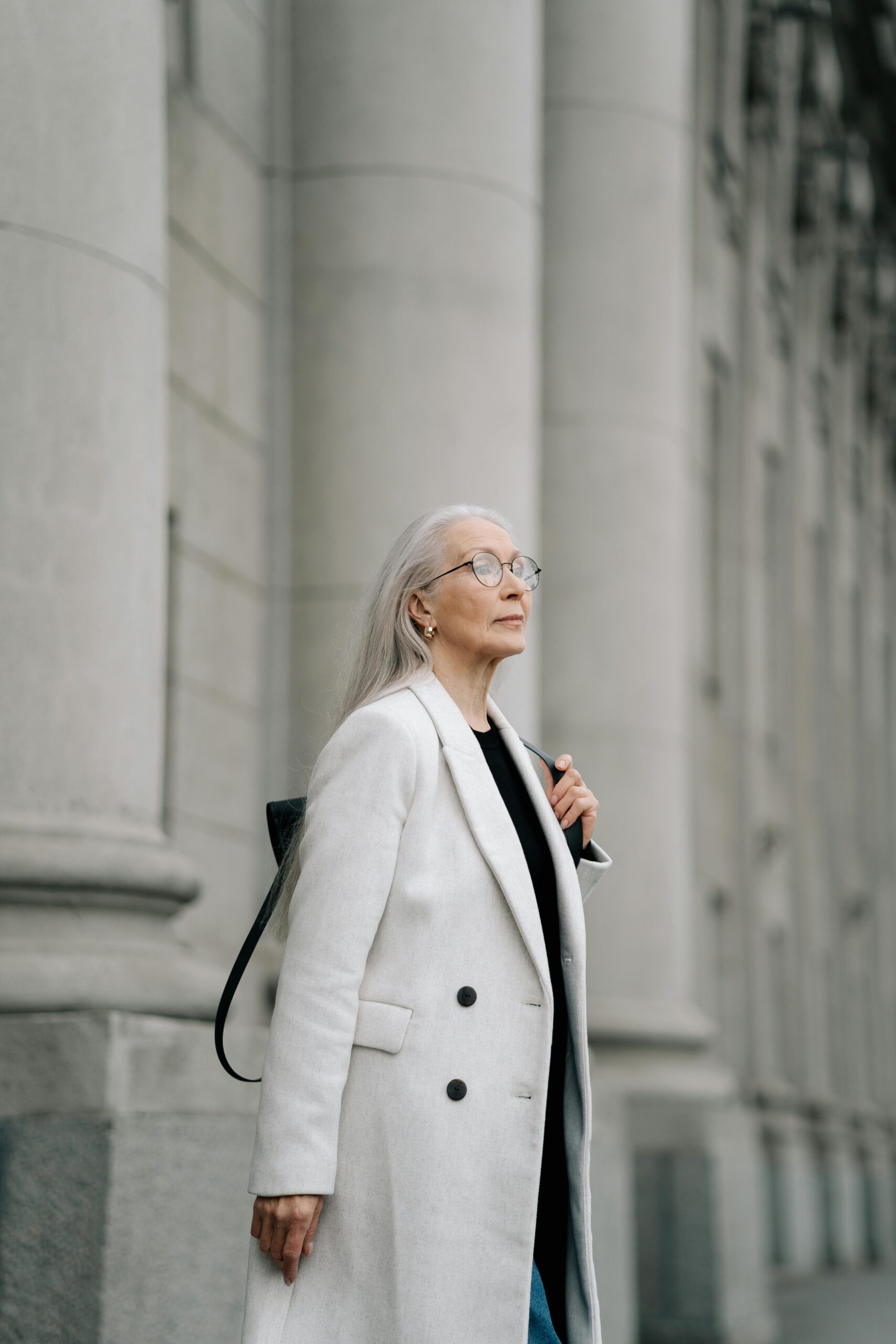 Senior woman with long gray hair and glasses in a white coat standing by columns.