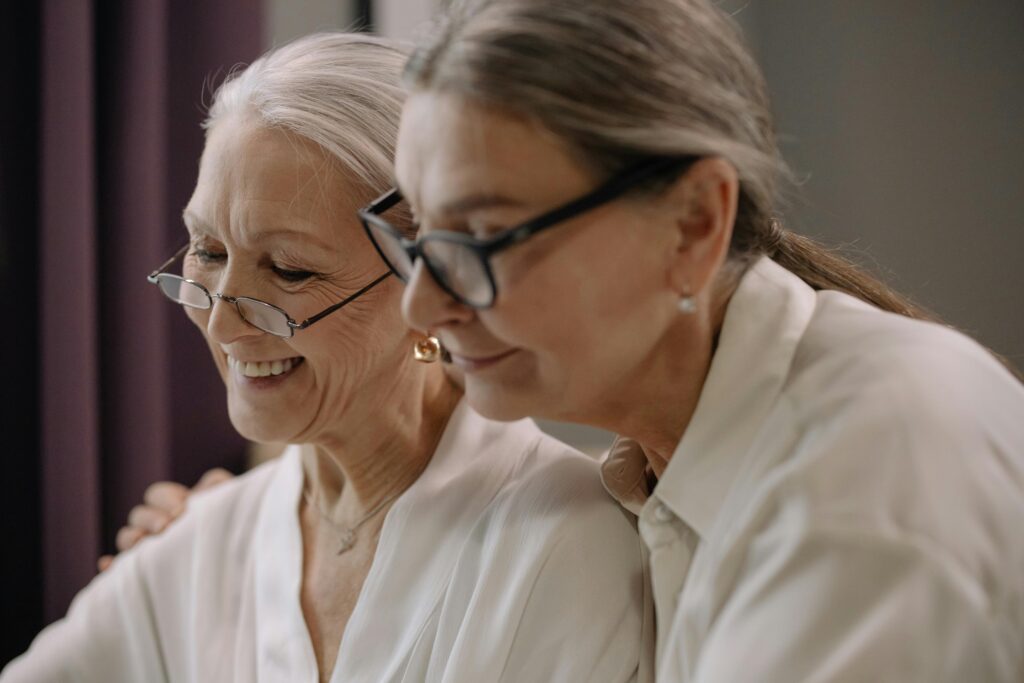 Two elderly women wearing glasses share a joyful moment indoors.