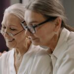 Two elderly women wearing glasses share a joyful moment indoors.